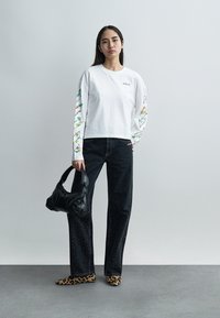 Γυναίκα στέκεται σε γκρίζο τοίχο φορώντας λευκό μακρυμάνικο πουκάμισο με φλοράλ μανίκια, μαύρα τζιν με πουά, παπούτσια λεοπάρ και κρατώντας μαύρο τσάντα.