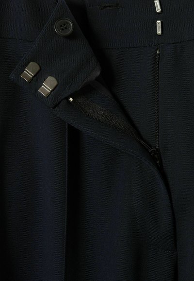 Primo piano di un tessuto blu navy che mostra una tasca con zip, dettagli con bottoni e bordi cuciti, evidenziando la texture e le caratteristiche di design.