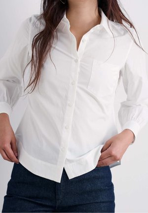 Donna che indossa una camicetta bianca con bottoni e taschino sul petto e jeans blu scuro, che sistema l'orlo da un lato.