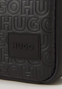 Crna teksturirana kožna površina s utisnutim ponavljajućim natpisom "HUGO" i središnjom šavom sa uzdignutim logom "HUGO".