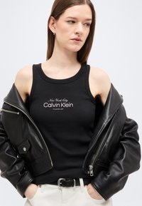 Γυναίκα που φορεί μαύρο κορυφαίο τοπ Calvin Klein, άσπρο παντελόνι και μερικώς αφαιρεμένο μαύρο δερμάτινο μπουφάν, κοιτάζοντας προς τα πλάγια.