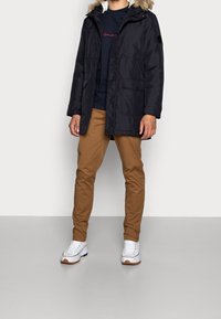 Marinkappa med fuskpälsfodrad huva, dragkedja och knappstängning, två framfickor; kombinerad med beiga chinos och vita sneakers med bruna detaljer.