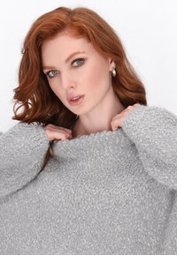Grauer, flauschiger Pullover mit strukturierter Oberfläche, weitem Rundhalsausschnitt und lockerer Passform. Das Model zieht am Kragen und präsentiert das weiche Material und den entspannten Stil.