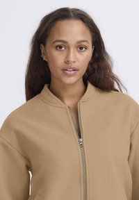 Tamn zip-up jakna s mekanim, teksturiranim materijalom, koja ima visoki ovratnik i opušteni kroj, prikazana na svijetloj pozadini.
