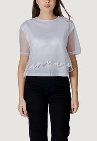 Camiseta blanca cortada con mangas de malla transparente, que presenta el bordado "Armani" en el dobladillo; textura suave y corte relajado. Llevada con pantalones negros.