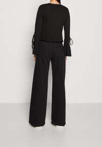 Sort langærmet top med flagrende ærmer og snøredetaljer, parret med vidde sorte jeans og hvide sneakers. Glat stofstruktur.