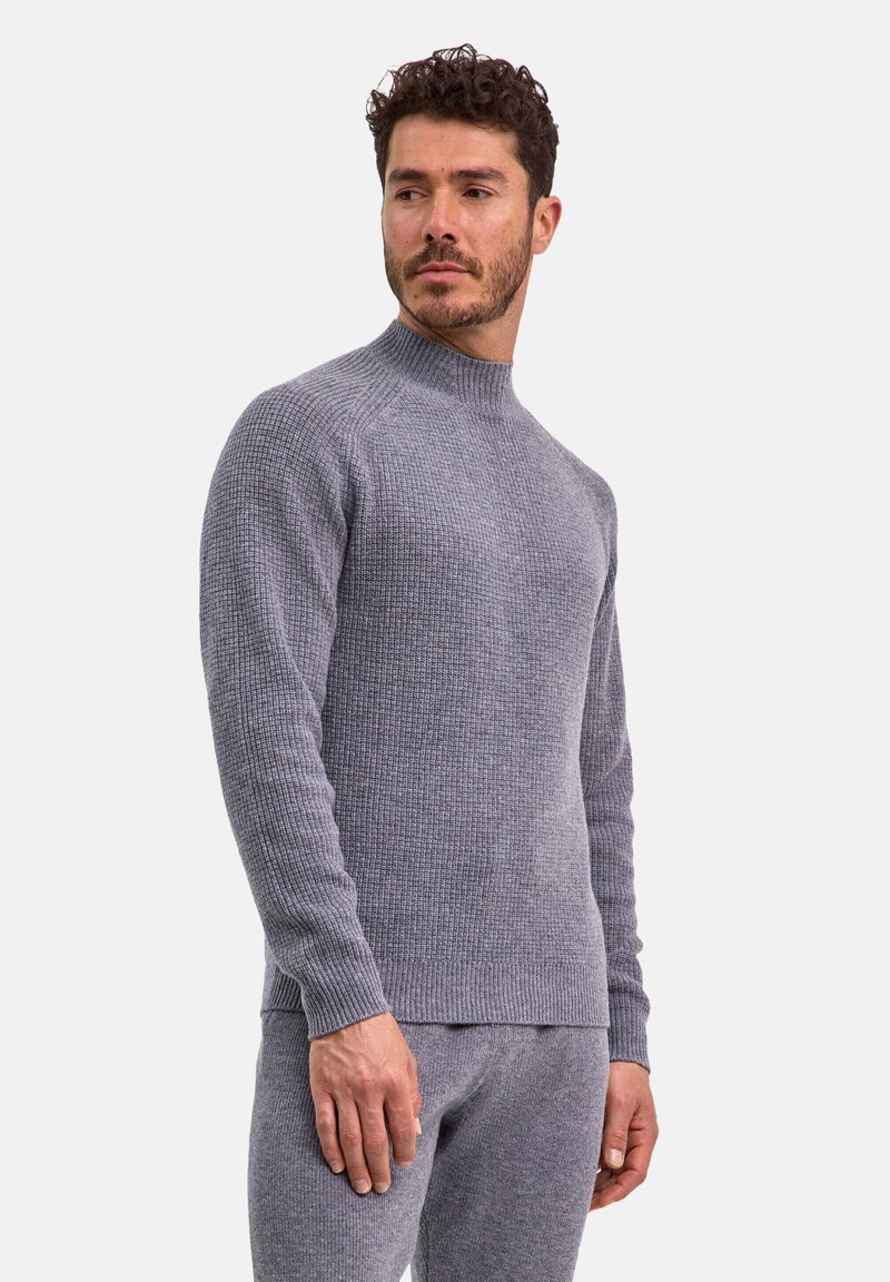 Grauer Strickpullover mit hohem Kragen, geripptem Muster und langen Ärmeln, kombiniert mit passenden grauen Hosen, die ein tailliertes Design zeigen.