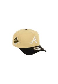 Beige baseball cap with a black bill, white embroidered "A" logo on the front, and a gold and black patch on the side.