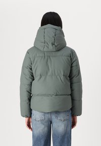 Padded jacket mutedinės žalios spalvos su kapišonu, su horizontaliomis siūlėmis ir laisvu kirpimu. Derinama su šviesiai mėlynomis džinsomis.