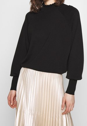 Vrouw draagt een zwarte trui met lange mouwen en een gerimpelde halslijn, gecombineerd met een glanzende geplooide crèmekleurige rok, staand tegen een effen achtergrond.