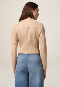 Giacca cropped in pelle beige con maniche lunghe, cuciture piatte e design minimalista, abbinata a jeans azzurri a vita alta.
