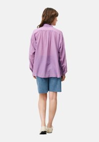 Lichtpaarse shirt met een relaxte pasvorm, gathered rug en lange mouwen; gecombineerd met denim shorts en beige instapschoenen.