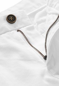 Pantalons en coton blanc avec un bouton en bronze et une fermeture éclair. La fermeture éclair présente des dents métalliques brunes contrastantes, accompagnées d'une texture de tissu lisse.