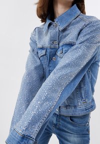 Nainen, joka pitää päällään vaaleansinistä farkkutakkia ja farkkuja, takki on koristeltu tiheästi sijoitetuilla pienillä hopeasoluilla hihoissa ja ylärinnassa.