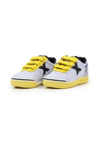 Zapatillas deportivas blancas y amarillas con diseño negro en forma de 'X', tres correas de velcro amarillas y un talón estampado, exhibidas una al lado de la otra sobre un fondo blanco.