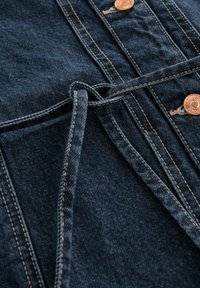 Tumma sininen farkkukangas, jossa on näkyvät ompeleet, solmittu vyö ja kupariset napit, jolla on sileä tekstuuri ja jäsennelty muotoilu.