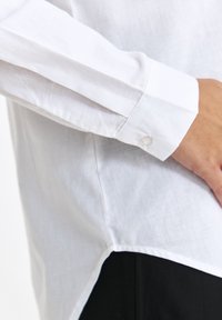 Biała lniana koszula z długimi rękawami, zapinanym mankietem i zaokrąglonym dołem. Czarny akcent widoczny na dolnej krawędzi, podkreślający kontrast kolorów.