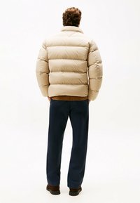 Beżowa ocieplana kurtka z wysokim kołnierzem i poziomymi przeszyciami, zestawiona z ciemnoniebieskimi spodniami i brązowymi butami.