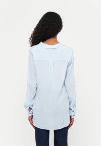 Light blue striped long-sleeve shirt with a rounded hem, back pleat, and button detail on sleeves. Soft fabric with a smooth texture.