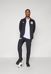 Fekete tréningruha piros részletekkel, mellkason emblémával, fehér pólóval és fekete futballcipővel párosítva. Egy futballon áll.