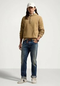 Sweatshirt bege com bolso frontal e cordões, jeans azuis em ganga e ténis brancos. Acessórios incluem um boné branco e um relógio.