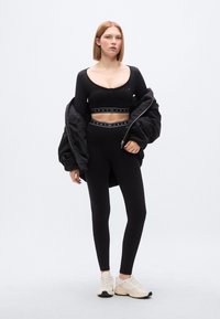 Μαύρο σετ αθλητικών ρούχων με κοντό μακρυμάνικο τοπ και κολάν υψηλής μέσης με ελαστικές ταινίες με λογότυπο. Το μοντέλο φοράει μαύρο πανωφόρι τύπου μπουφάν.