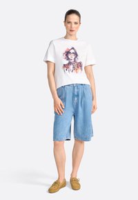 Tricou alb din bumbac cu un design grafic colorat ce prezintă un portret de femeie, asortat cu pantaloni scurți din blug albastri și pantofi din suede maro.