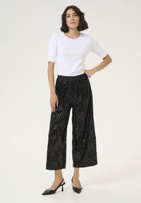 Pantaloni wide-leg neri con paillettes e vita elastica, abbinati a una blusa bianca aderente. La modella indossa scarpe con tacco a punta nera.