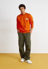 Oranža kapuce ar priekšējo logo, pārī ar olīvzaļām cargo biksēm ar sānu kabatām. Modelis uzvelk baltas sporta kurpes uz bēša grīdas.