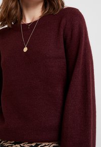 Pull tricoté bordeaux avec une texture côtelée et un large col rond, associé à un bas à motifs et des colliers superposés.