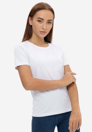 Giovane donna con capelli castani lisci che indossa una t-shirt bianca semplice a maniche corte e jeans blu su sfondo bianco.