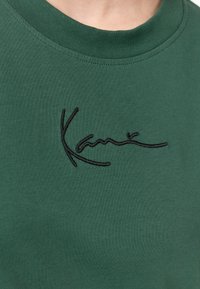T-shirt en coton vert à col rond. Présente un logo en texte noir brodé sur le devant, une texture lisse et des manches courtes.
