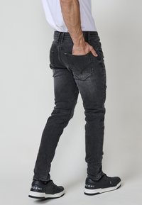Fekete, slim-fit farmernadrág, megkopott textúrával, oldalsó és hátsó zsebekkel. Fekete sportcipővel párosítva, fehér részletekkel.