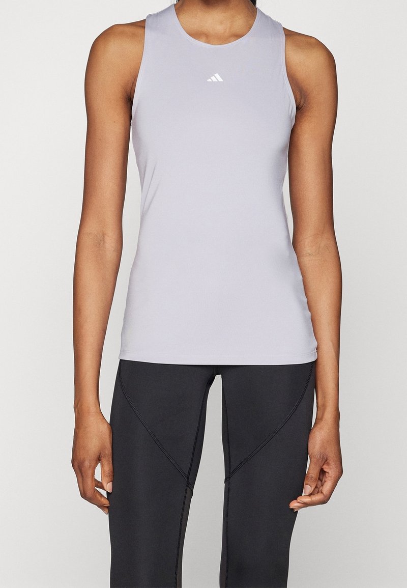 Světlé šedé bezrukávové sportovní triko z hladké látky, s kulatým výstřihem a malým bílým logem. Kombinováno s černými přiléhavými legínami.