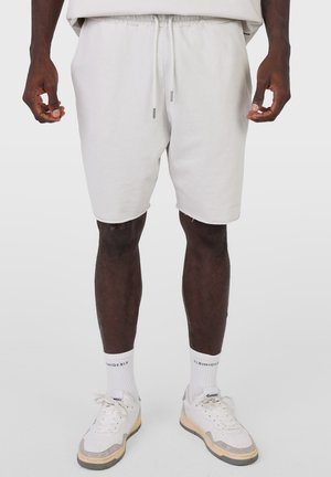 Mężczyzna noszący jasnoszorty ze sznurkiem, białe skarpety za kostkę z czarnym napisem i białe trampki z beżową podeszwą.