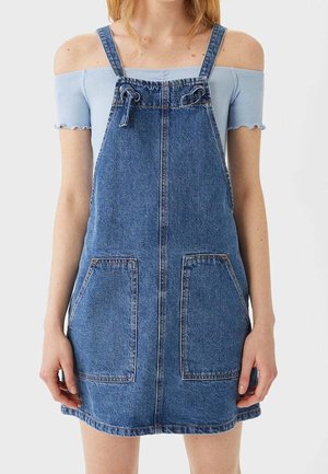 Kvinde iført en ærmeløs denim-selvbindende kjole over en lyseblå, off-shoulder, ribstrikket top, stående mod en ensfarvet lys baggrund.