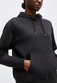 Άτομο που φοράει μαύρο φούτερ Puma Sportswear με μπροστινή τσέπη και κορδόνια, χέρι στην τσέπη, περικομμένο στον λαιμό και τους γοφούς.