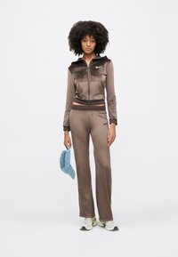 Γυναίκα με σγουρά μαλλιά που φορά καφέ βελουτέ φόρμα και λευκά αθλητικά παπούτσια, κρατώντας μπλε καπέλο μπέιζμπολ πάνω σε λευκό φόντο.