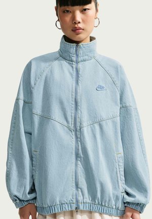 Junge Frau trägt eine hellblaue, übergroße Nike-Reißverschlussjacke mit hohem Kragen und Creolen, steht vor einem einfarbigen Hintergrund.