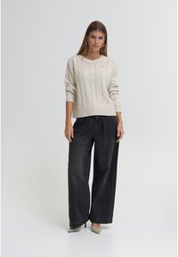 Krem pulover iz grobega pletenja, kombiniran s širokimi temnimi kavbojkami in bež petami. Model stoji na nevtralnem ozadju.