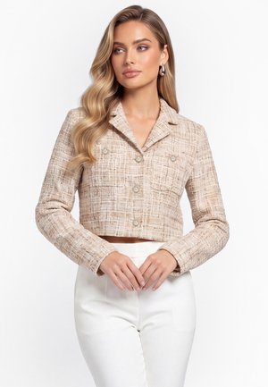 Vrouw met lang golvend blond haar draagt een beige gestructureerd cropped jasje met voorzakken en witte hoge taille broek, staand tegen een witte achtergrond.