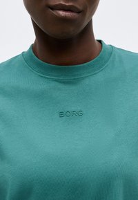 Κοντινό πλάνο ενός ατόμου που φοράει μια βεραμάν μπλουζάκι με στρογγυλή λαιμόκοψη και ανάγλυφη επιγραφή "BORG" στο στήθος.