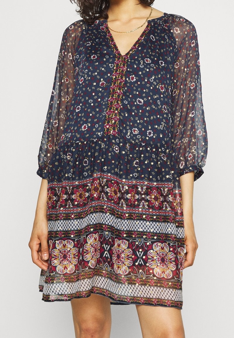 Femme portant une robe fleurie bleu marine longueur genou avec manches longues en voile et détails multicolores à motifs à l'ourlet et à l'encolure.