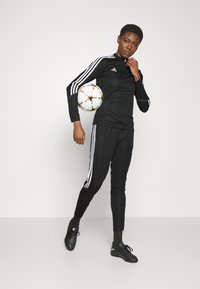 Čierny atletický outfit s bielymi pruhmi, obsahujúci tričko s dlhým rukávom a tesné nohavice. Súčasťou sú čierne futbalové kopa čky a viacfarebný futbalový lopt. 