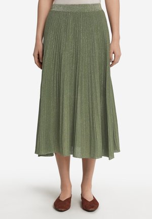 Jupe midi plissée en vert clair avec une subtile brillance. Taille élastique, forme fluide et texture lisse. Chaussures en ballerines brunes.