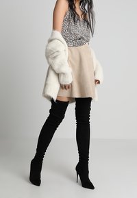 Cárdigan de piel sintética blanca sobre una blusa de leopardo, falda beige y botas de muslo negras de ante con tacones de aguja.