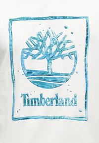 Μπλε λογότυπο Timberland με ένα στυλιζαρισμένο δέντρο μέσα σε κύκλο και το όνομα της μάρκας από κάτω, όλα περιτριγυρισμένα από ένα μπλε ορθογώνιο πλαίσιο.