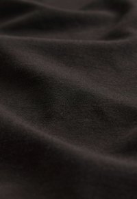 Musta puuvillakangas, jonka pinta on sileä ja jossa on hienovaraisia taitoksia ja ryppyjä, esittelee syvää mattaista viimeistelyä.