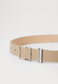 Beige kožený opasek s hladkým povrchem, stříbrnou kovovou sponou a nastavitelnými otvory pro přizpůsobení velikosti. Detail loga na sponě.
