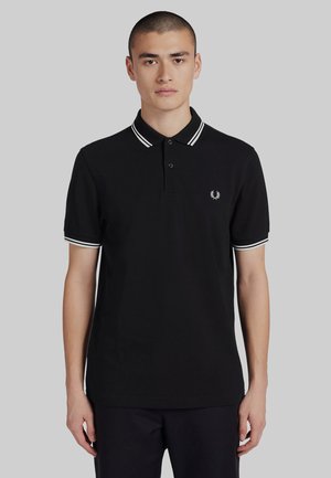 Joven con cabello corto que lleva una camiseta polo negra con el cuello y los bordes de las mangas a rayas blancas, posando frente a un fondo gris liso.